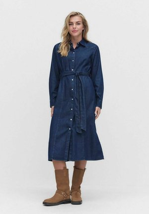 Kvinna i en långärmad, knäppt klänning av mörk denim med ett midjeband och bruna stövlar som går till vaden, stående framför en enfärgad bakgrund.