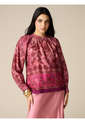 Donna con capelli lunghi e scuri che indossa una camicetta rosa a motivo floreale con maniche lunghe e una gonna rosa chiaro su uno sfondo beige.