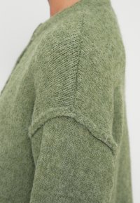 Πλεκτό πουλόβερ σε απαλό πράσινο μαλλί με υφή και φινίρισμα, χαρακτηρισμένο από στρογγυλή λαιμόκοψη και σχεδιασμό με χαμηλωμένους ώμους.