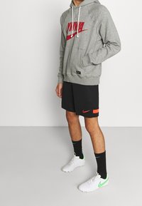 Personne portant un sweat à capuche Nike gris avec logo rouge, un short Nike noir avec détails rouges, des chaussettes noires, et des crampons Nike blancs avec le swoosh vert.