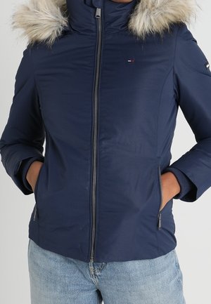 Personne portant une veste d'hiver bleu marine avec une fermeture éclair devant, une capuche bordée de fourrure et les mains dans les poches latérales, associée à un jean bleu clair.