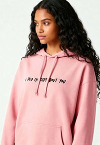 Felpa rosa realizzata in materiale morbido, con tasca frontale e testo nero che recita "HO PARLATO DI TE A CHATGPT." Design casual.