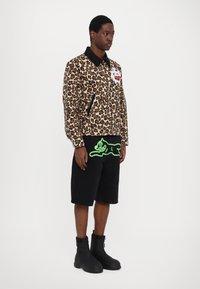 Giacca con stampa leopardata e colletto nero, tasche con zip e grafica bianca. Pantaloni corti neri con grafica di un gatto verde neon. Stivali neri.