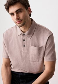 Lysebrune kortærmede polo shirt med flad krave, tre-knap lukning og brystlomme. Stoffet har en blød tekstur.