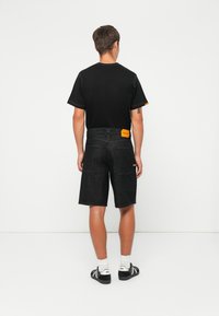 Man wearing black t-shirt, dark denim shorts with orange patches, white socks, and black sneakers standing against a plain white background.
