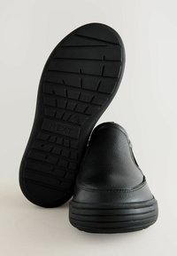 Chaussure noire à enfiler avec un dessus en cuir texturé, semelle en caoutchouc dotée d'un motif de semelle pour l'adhérence, et logo embossé sur le côté.