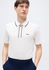 Λευκό πόλο μπλουζάκι με φερμουάρ στη μέση της γλώσσας, μαύρες και μπεζ λωρίδες διακόσμησης, και το λογότυπο "BOSS" στην αριστερή πλευρά. Κοντά μανίκια και απαλή υφή.