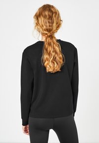 Svart sweatshirt med långa ärmar och rund hals, tillverkad av mjukt material, visas bakifrån, i kombination med svarta leggings.