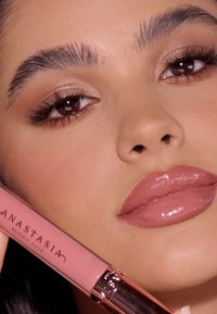 Roze lipproduct in een heldere tube met een metallic roségouden dop, gehouden naast een gezicht met opvallende lippen, gedefinieerde wenkbrauwen en een natuurlijke make-up.