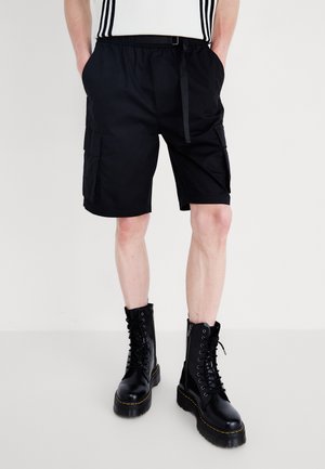 Personne portant un short cargo noir avec des poches et des bottes de combat noires à lacets, debout devant un fond uni.