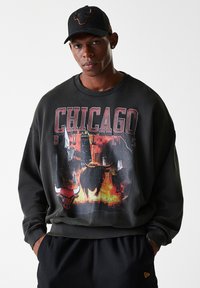 Sweat-shirt noir avec un graphique des Chicago Bulls représentant un taureau et la silhouette de la ville. Accompagné d'une casquette noire avec le logo d'un taureau.
