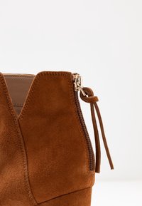 Ruskean mokkanahkasaappaat nilkkapituiset, joissa on takana vetskari ja nahkakahva, vaaleaa taustaa vasten.