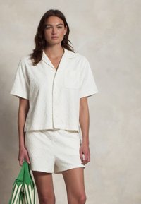 Chemise blanche texturée à manches courtes avec un short assorti. Dotée d'une poche, d'un motif géométrique et d'une coupe décontractée. Sac rayé vert à la main.