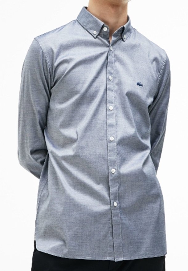 Homme portant une chemise bleu clair à boutons avec col boutonné et petit logo brodé sur le côté gauche de la poitrine.