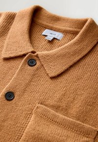 Braun strukturierte Strickkragenhemd mit einer Brusttasche, ausgestattet mit zwei schwarzen Knöpfen und einem Etikett mit der Aufschrift "NEXT" unter dem Kragen.