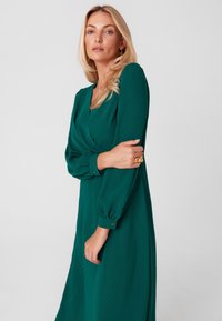 Zielona sukienka w kształcie kopertowym, wykonana z gładkiego materiału, z długimi bufkowymi rękawami i zapinanymi mankietami. Design obejmuje głęboki dekolt w kształcie litery V.