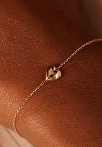 Gouden hartvormige armband op gestructureerde bruine huid, met een delicate ketting die aansluit op de gepolijste hartbedel.