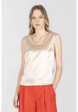 Kobieta ubrana w bezrękawnikowy, satynowy top w kolorze szampańskim z kryształkowym dekoltem oraz plisowane pomarańczowe spodnie z wysokim stanem na białym tle.