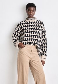 Strickpullover in Creme und Schwarz mit geometrischem Muster, hohem Kragen und lässiger Passform. Kombiniert mit hellbeigen Cordhosen.