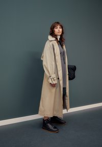 Beige trench coat, lång och lös passform, med knäppta ärmslut. Bärs över en grå klänning, kombinerad med svarta hög-top sneakers, stående mot en grön bakgrund.
