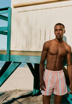 Shirtloser Mann in rosa-weiß gestreiften Badeshorts steht neben einem türkis-beigen Rettungsturm am Sandstrand.