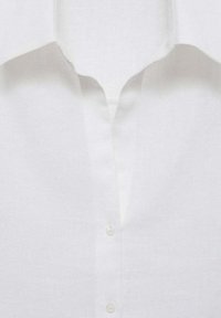 Devant d’une chemise en lin blanc avec un col cranté et deux boutons visibles sur un fond de tissu lisse.