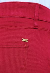 Vaqueros de mezclilla rojos con una pequeña etiqueta dorada "HILFIGER" en el bolsillo trasero, con patrones de costura visibles y una textura suave.