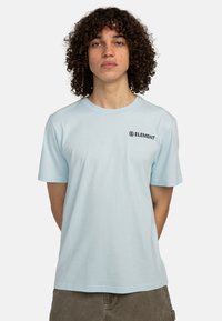 Light blue cotton t-shirt, short sleeves, crew neck. Features a black logo on the left chest. Smooth texture, relaxed fit.