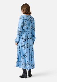 Long-sleeve blue dress with black leaf print, features a gathered back, belted waist, and flowy fabric, paired with black ankle boots.