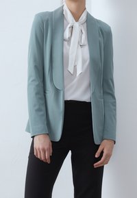 Blazer bleu clair à coupe ajustée, tissu lisse et revers à crans. Porté sur un chemisier blanc avec un nœud papillon. Associé à un pantalon noir.