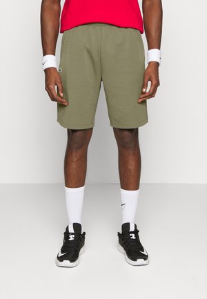 Mann trägt olivgrüne Shorts, weiße Nike-Handgelenksbänder und Socken, schwarz-weiße Nike-Sneaker, steht vor einem schlichten hellen Hintergrund.