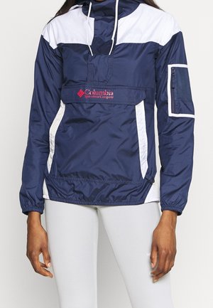 Persoon draagt een marineblauwe en witte Columbia Sportswear windbreaker met voorvak en witte trekkoorden, gecombineerd met lichtgrijze broek.