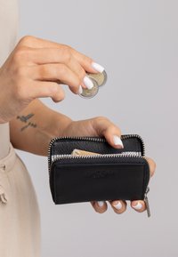 Schwarze Leder-Geldbörse mit silbernen Beschlägen, Reißverschluss und Innenfach. Hand hält Münzen über der Geldbörse.