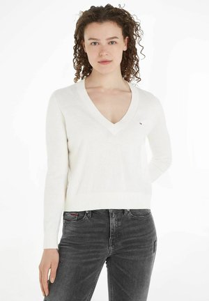 Maglione bianco leggero a collo a V con maniche lunghe, polsini e orlo a costine, con un piccolo logo del marchio sul petto. Abbinato a jeans scuri.