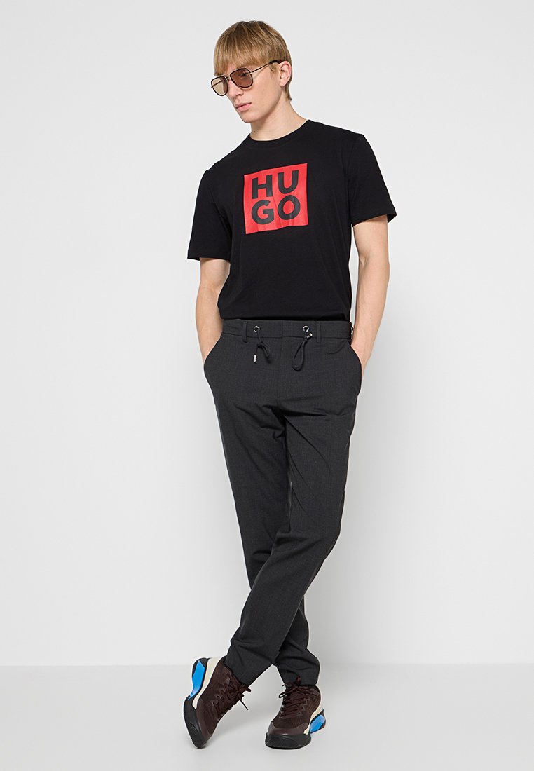 Camiseta negra con logo rojo "HUGO", pantalones de chándal grises con cordón y zapatillas deportivas marrones con acentos azules. Diseño minimalista y estilo ajustado.