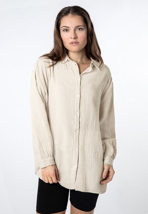 Light beige long-sleeve button-up shirt with a textured fabric, front buttons, and a collar. Paired with black shorts.