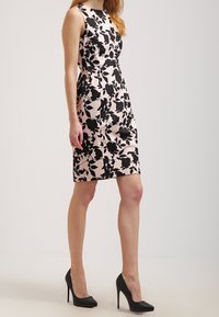 Robe à imprimé floral noir et blanc dans une silhouette ajustée, avec un design sans manches et un ourlet au genou, associée à des escarpins noirs.