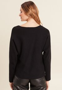 Schwarzer Strickpullover mit weitem Bootsausschnitt und langen Ärmeln, der einen lockeren Sitz und ein geripptes Material aufweist. Sichtbare Nahtdetails am Rücken.