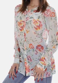 Blusa floreale realizzata in tessuto trasparente, con uno sfondo azzurro chiaro e fiori rosa e arancioni, maniche lunghe e chiusura con bottoni.