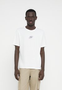 Camiseta blanca de algodón con un pequeño parche de logo en tonos púrpura y azul en el pecho. Mangas cortas, cuello redondo, corte regular. Diseño informal.