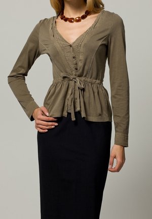 Oliwkowa bluzka z długim rękawem, dekoltem w serek, ozdobiona falbanką i wiązana w talii. Do niej dopasowana, czarna spódnica o gładkiej fakturze.
