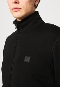 Black zip-up jacket made of soft fabric, featuring a high collar, contrasting stitching, and a small embossed logo on the chest.