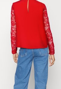 Donna che indossa un top rosso con maniche lunghe in pizzo trasparente a fiori e jeans a vita alta blu, vista di spalle su uno sfondo bianco.