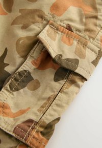 Tessuto mimetico caratterizzato da una combinazione di forme beige, marrone e verde. Include una tasca con patta e dettagli di cucitura a vista.