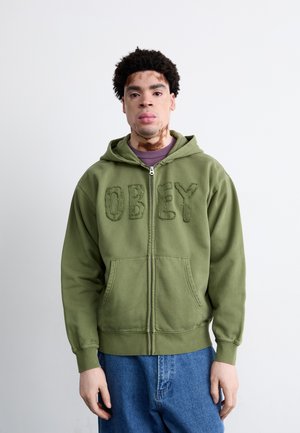Grüne Zip-Hoodie mit strukturiertem "OBEY"-Schriftzug, Fronttaschen und Kapuze mit Kordelzug. Getragen mit lockeren blauen Jeans.
