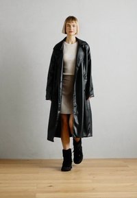 Fekete, fényes esőkabát vállra húzható epaulettekkel, dupla gombokkal, világos bézs ruhára és fekete bokacsizmákra húzva.