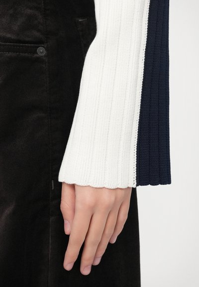 Manga de suéter de punto con franjas alternas blancas y azul marino, con textura acanalada y borde ondulado, combinada con un fondo de tela negra.