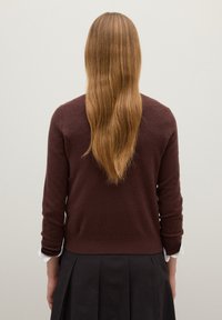 Maglione marrone scuro con collo a giro, maniche lunghe e orlo aderente, abbinato a una gonna nera plissettata. Capelli lunghi e lisci, di colore marrone.