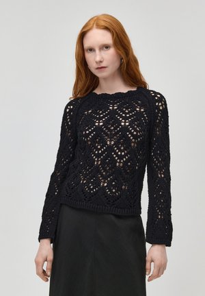 Žena s červenými vlasy, oblečená v černém dlouhém svetr z otevřeného úpletu a černé sukni, stojící před jednoduchým světlým pozadím.