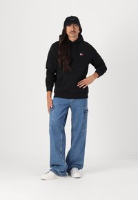 Schwarzer Kapuzenpullover mit Fronttasche, kombiniert mit lockeren blauen Denim-Jeans. Weiße Sneakers vervollständigen das Outfit. Die Mütze ist schwarz mit Logo.